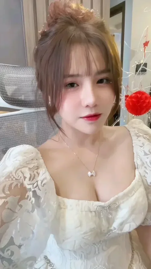 Trong khung hình phòng ngủ, cô gái tóc buộc cao mặc váy ren trắng, vú tròn đầy lộ nhẹ